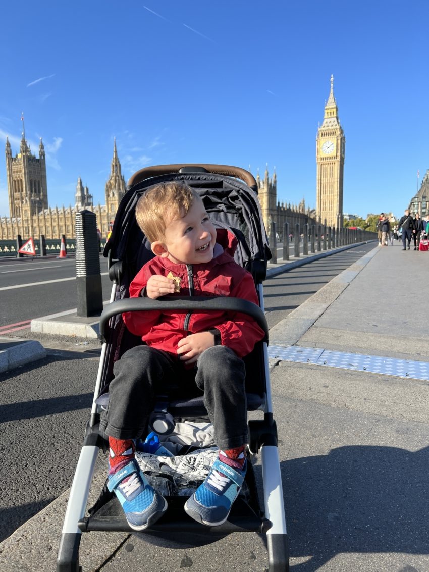 Smiling toddler in a red jacket sitting in a compact stroller suitable for airplane travel, with the iconic Big Ben and the Houses of Parliament in the background, epitomizing joyful family adventures in London.